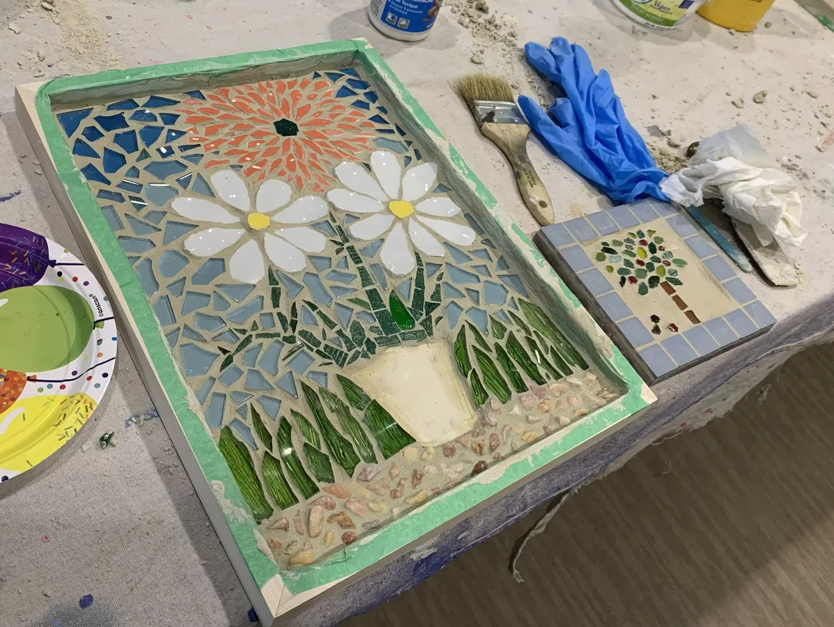 An unfinished mosaic piece of flowers in a flower pot on a table with gloves, paints and glue.