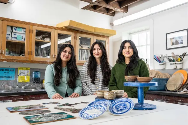 Three figures stand in a ceramic studio with their completed ceramic pieces in front of them on a table top.