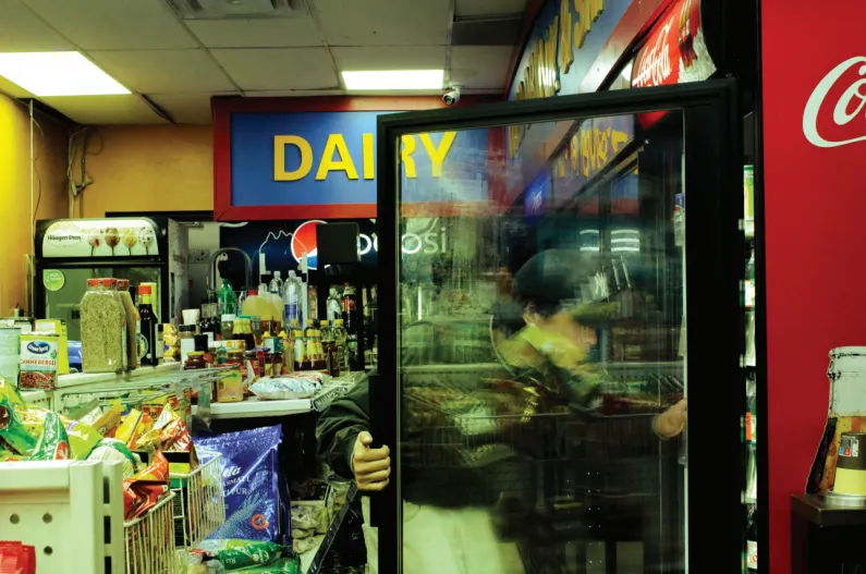 Someone opens a convenience store fridge door and looks inside, behind them are shelves full of snacks.